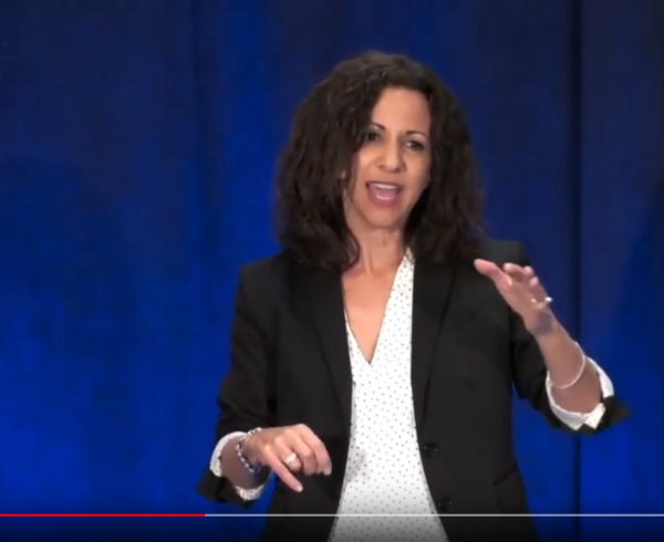 Woman speaking and gesturing on stage with blue background.