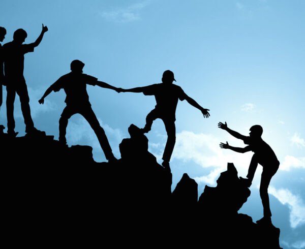 Silhouettes of people helping each other climb a rocky hill at sunset.